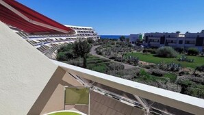 A balcony with a green table and chairs, overlooking a landscaped area with a pool and buildings in the background. - AMOUR AMOUR - Apartment Héliopolis K 209 - Luxe - 4 people (AGDE)