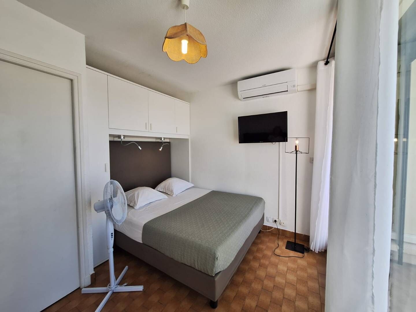 A bedroom with a bed, a standing fan, a mounted TV, and an air conditioning unit.