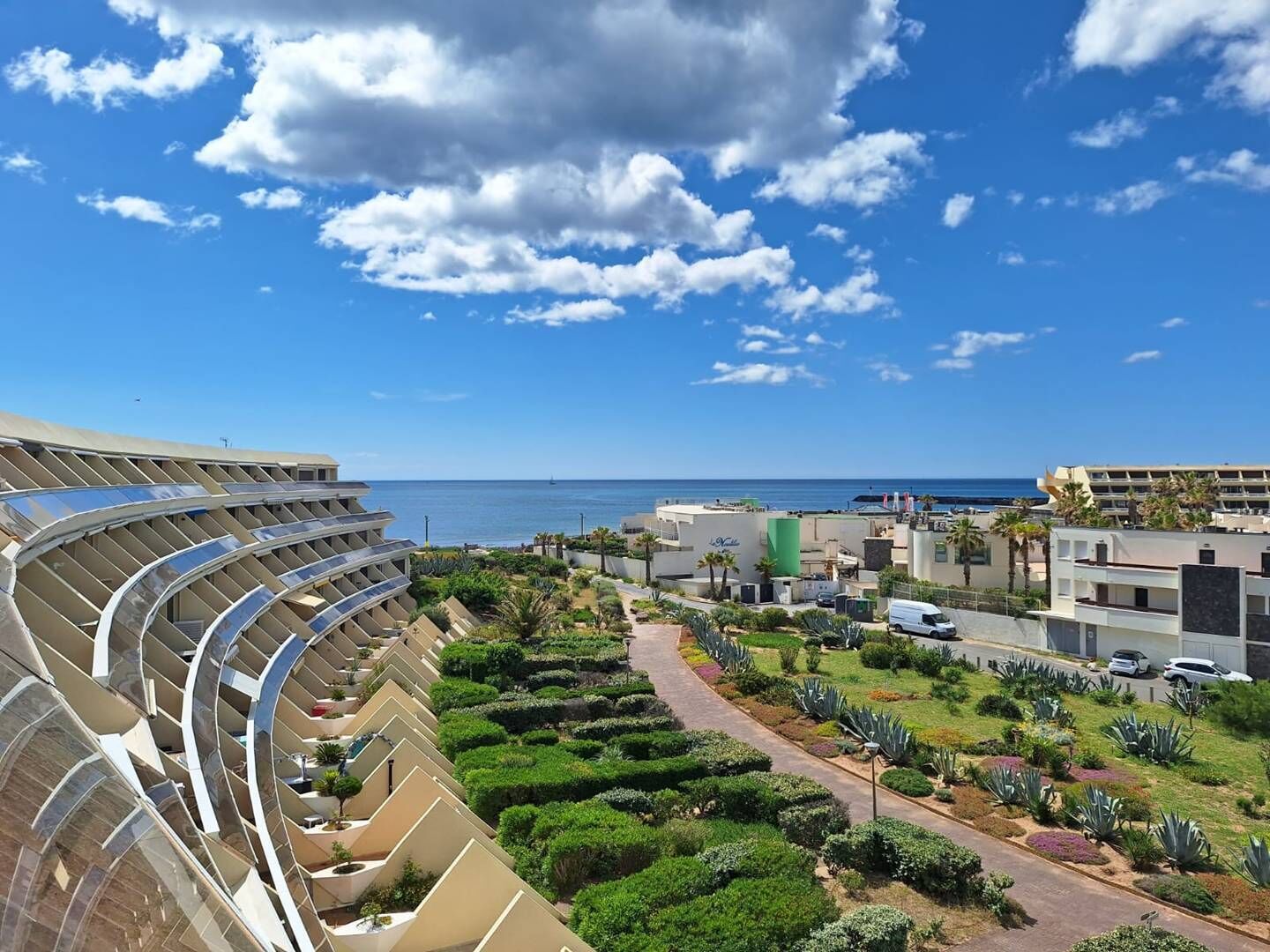 Un hôtel moderne doté d’un balcon aux lignes courbes, offrant une vue sur un jardin aménagé et une plage.