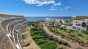 Een modern hotel met een balkon in een gebogen ontwerp, uitkijkend over een aangelegde tuin en een strand.