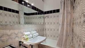 Een badkamer met een wit wastafel, een bad en een douchegedeelte. Er liggen handdoeken en toiletartikelen op de wastafel.