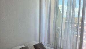 A single bed with white bedding and two pillows, a window with sheer curtains, and a view of outdoor furniture. - Studio Héliopolis L 20 - 2 people (AGDE)