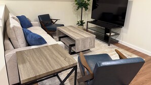 Un salon moderne avec un canapé d’angle, une table basse en bois, un fauteuil bleu, un téléviseur posé sur un meuble et une plante en pot.