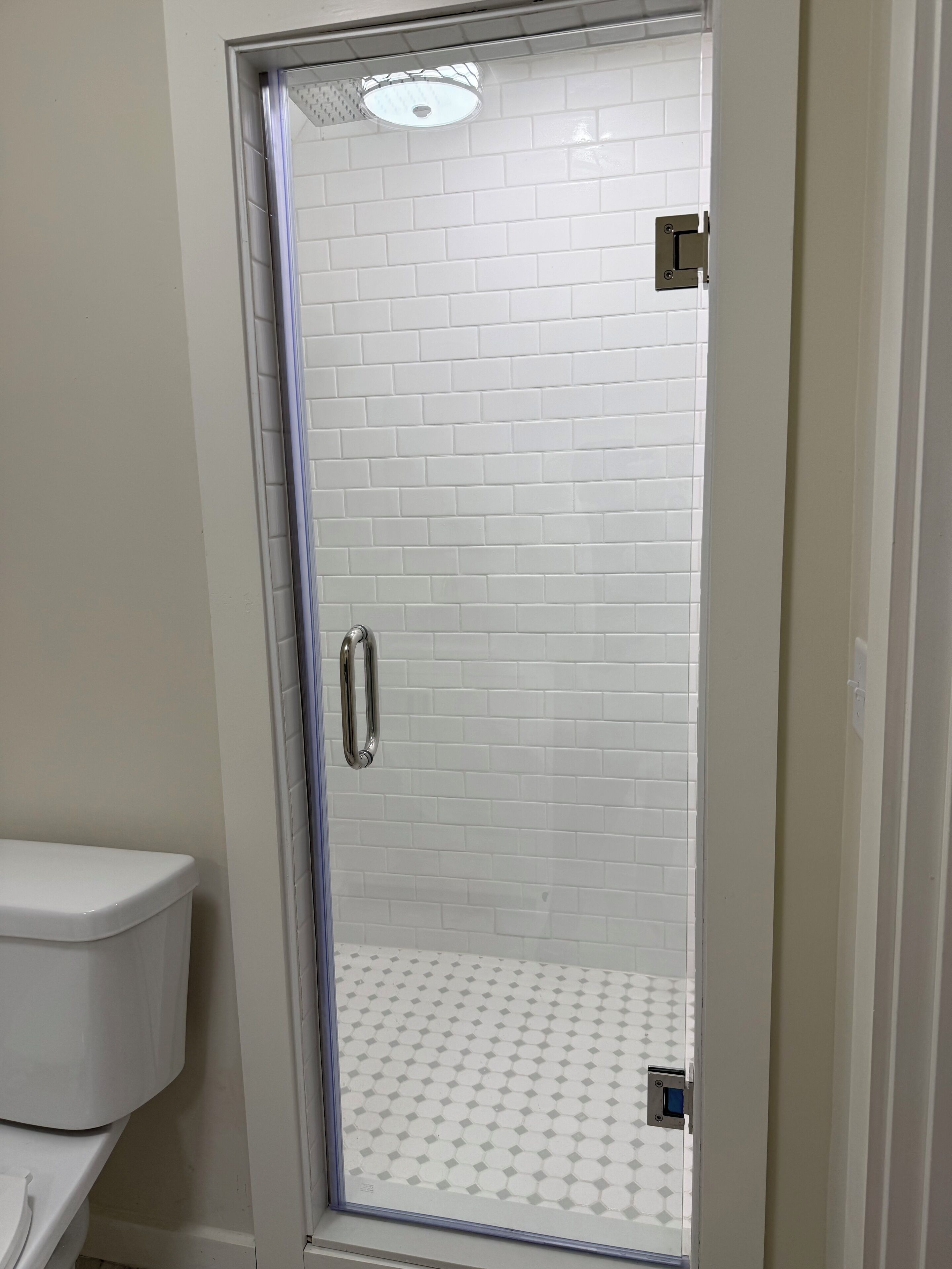 Une salle de bain avec des toilettes blanches, une douche à parois de verre et un mur recouvert de carreaux blancs, ainsi qu’un panneau de commande visible.