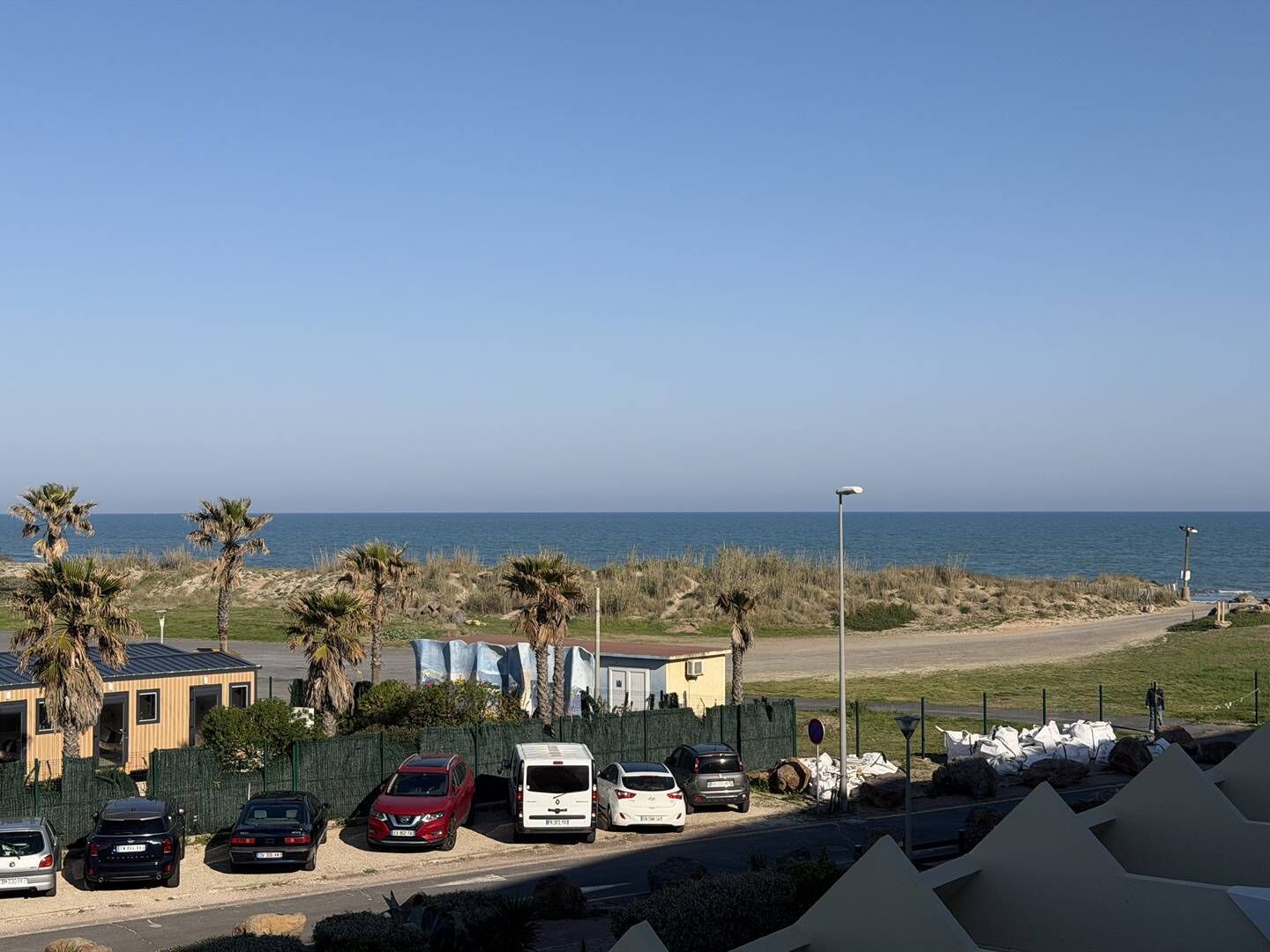 A parking area with several cars, a building with a sloped roof, and a view of the ocean.