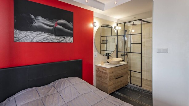 Ein modernes Schlafzimmer mit einem Bett, einer roten Akzentwand, einem großen Spiegel und einer Duschkabine.