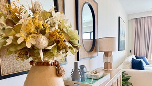 Uma sala de estar com um vaso grande de flores, um espelho, uma luminária e um sofá com almofadas.