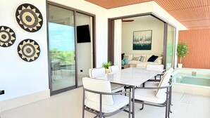 Uma área de refeições moderna ao ar livre, com mesa de vidro e cadeiras brancas, um sofá e um quadro na parede.