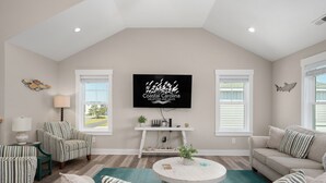 A living room with a high ceiling, a flat-screen TV mounted on the wall, a striped armchair, a sofa, a round coffee table, and a window with blinds. - Rock Me on the Water: Oceanside, Pool, Hot Tub (Nags Head)