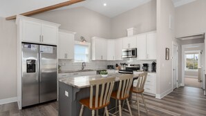 A modern kitchen with white cabinets, stainless steel appliances, a large island with bar stools, and a wooden ceiling beam. - Rock Me on the Water: Oceanside, Pool, Hot Tub (Nags Head)