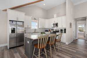 A modern kitchen with white cabinets, stainless steel appliances, a large island with bar stools, and a wooden ceiling beam. - Rock Me on the Water: Oceanside, Pool, Hot Tub (Nags Head)