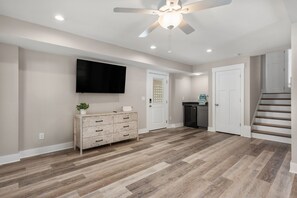A spacious room with a wooden floor, a ceiling fan, recessed lighting, a flat-screen TV, a wooden cabinet, and a staircase leading to another level. - Rock Me on the Water: Oceanside, Pool, Hot Tub (Nags Head)
