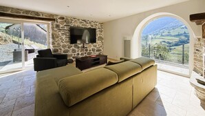Ein geräumiges Wohnzimmer mit Steinwand, einer großen Couch, einem Flachbildfernseher und Blick auf eine ländliche Landschaft durch ein Rundbogenfenster.
