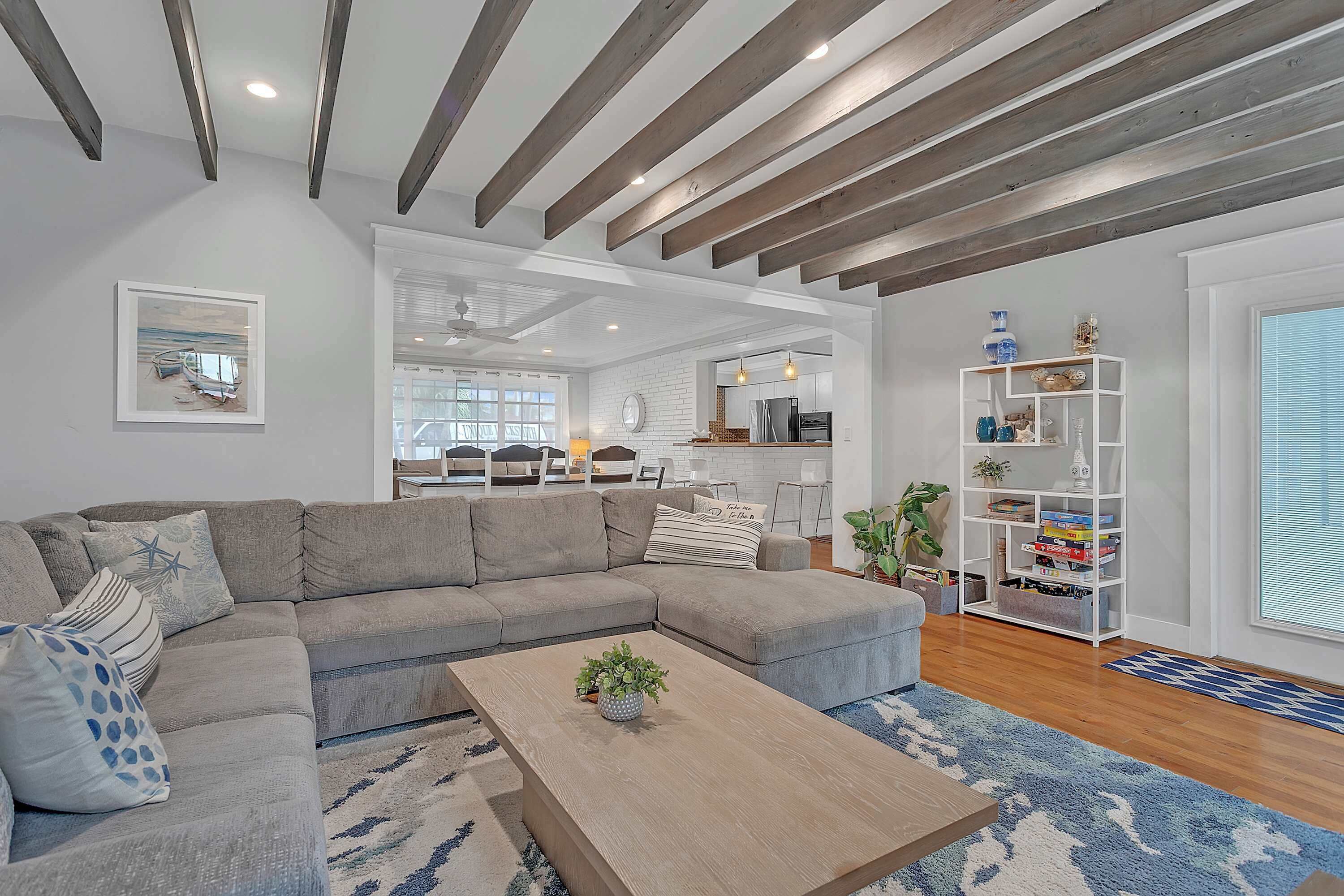 A modern living room with a sectional sofa, a coffee table with a plant, a white bookshelf, and a kitchen area in the background.