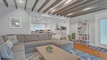 A modern living room with a sectional sofa, a coffee table with a plant, a white bookshelf, and a kitchen area in the background.
