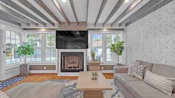 A living room with a fireplace, large windows, a flat-screen TV, and a sofa.