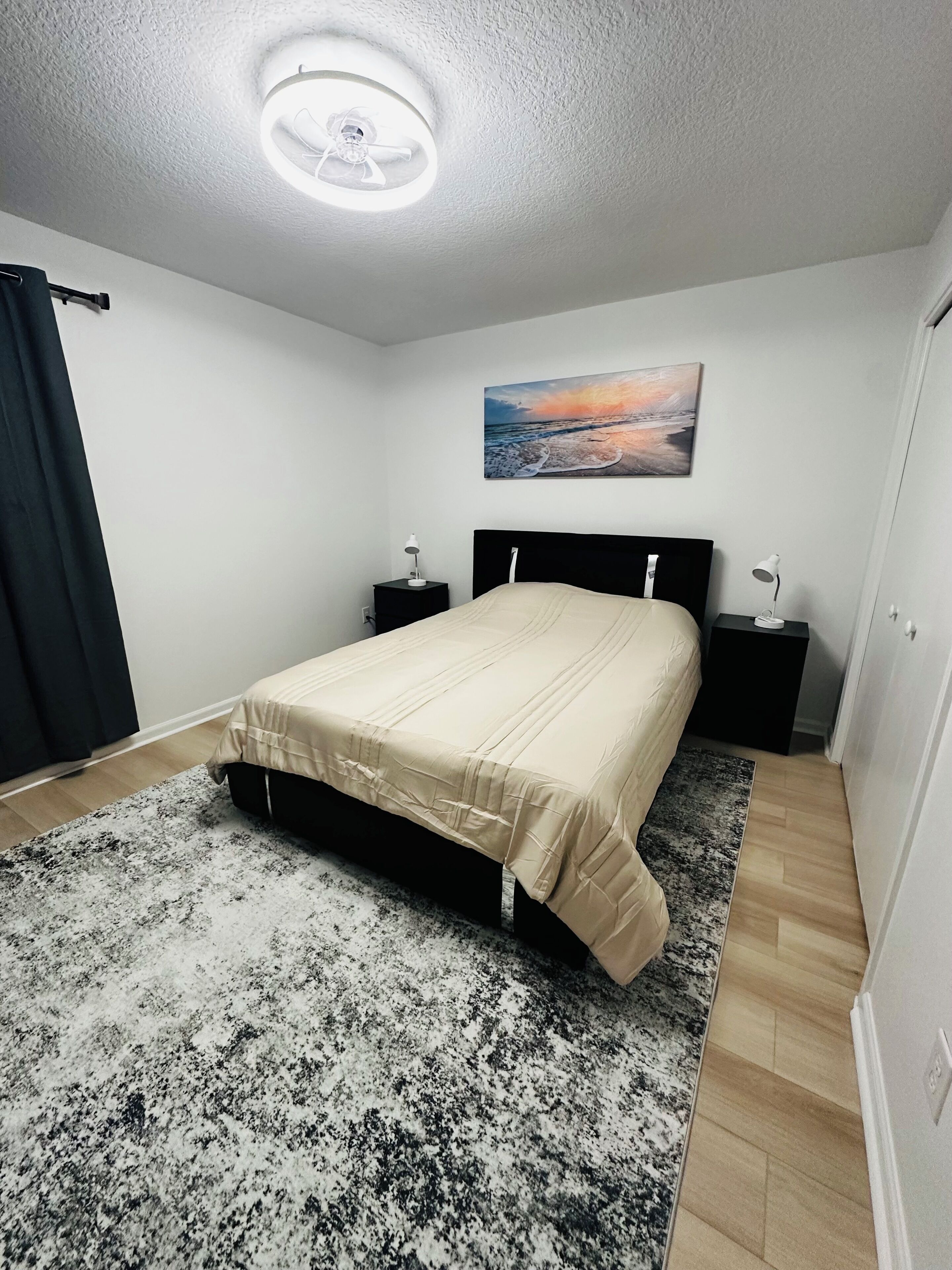 A bedroom with a bed, two bedside tables, a nightstand, a ceiling light, a painting, and a window with curtains.