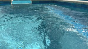 A swimming pool with clear blue water, a tiled edge, and a building with a brown roof and glass doors in the background. - Mary's Villas  (Oranjestad)