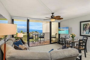 A living room with a sofa, a TV, a ceiling fan, and a balcony with a table and chairs. - Kaanapali Shores | Ocean View 4 BR, Sleeps 12 | Car Incl. w/ 6+ Nights | KS ML-3677 by KBM (Lahaina)
