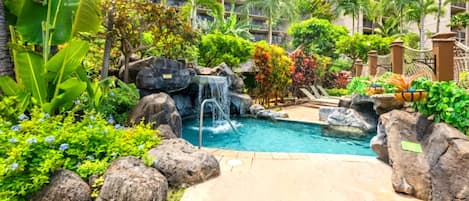 Zona de piscina con cascada, rodeada de plantas tropicales y palmeras, con un edificio al fondo.