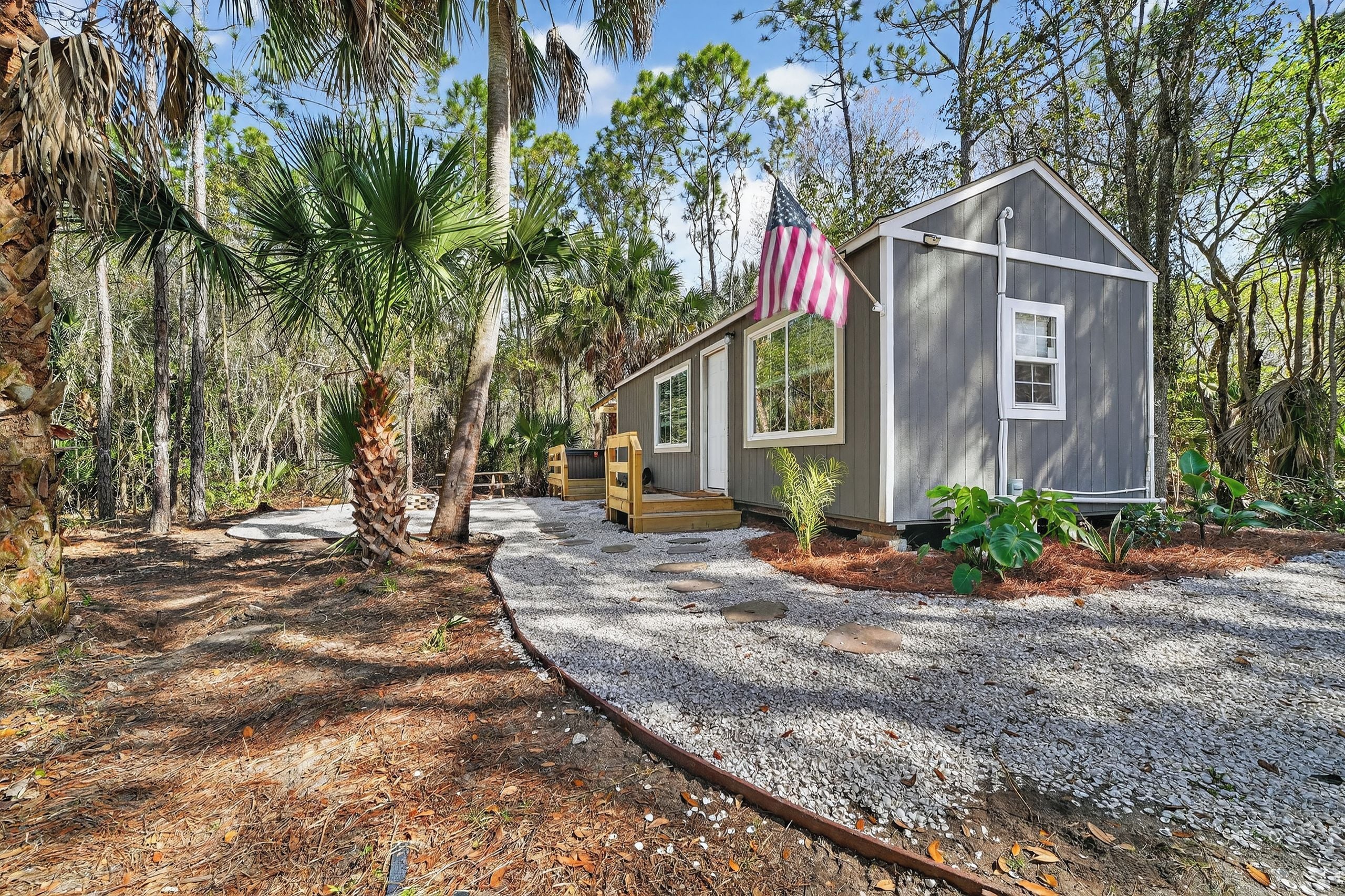 A small cabin with a gravel driveway, surrounded by trees and a flag.