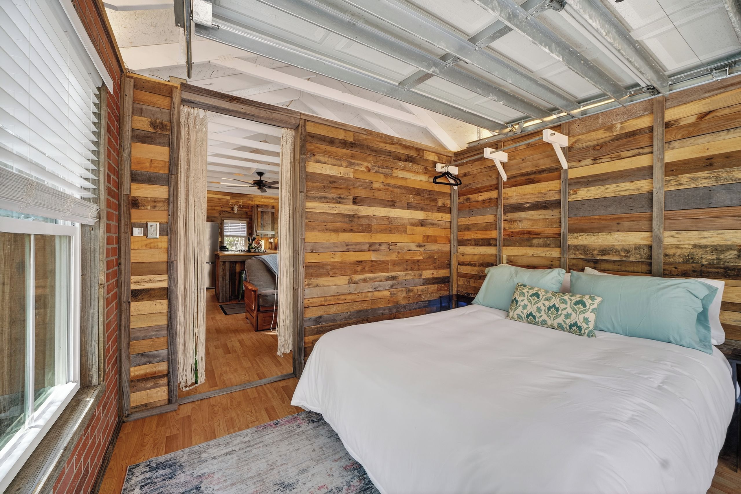 A bedroom with a wooden slat wall, a bed with white bedding, and a view of a kitchen and bathroom through an open door.