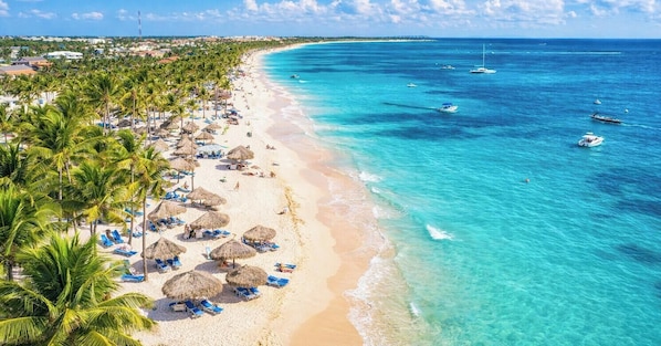 A beach with clear turquoise waters, palm trees, and beach umbrellas. - Modern Poolside Villa in Vistacana Near Downtown (Punta Cana)