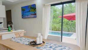 Ein Essbereich mit Holztisch, weißen Stühlen, einer blau-weißen Tischläufer und einem Kerzenarrangement. An der Wand hängt ein Gemälde vom tropischen Strand, und ein Fenster mit einem roten Sonnenschirm blickt auf einen Swimmingpool.