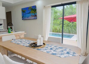 Un comedor con una mesa de madera, sillas blancas, un camino de mesa azul y blanco estampado y un centro de mesa con velas. Un cuadro de una playa tropical adorna la pared, y una ventana con un paraguas rojo da vista a una piscina.