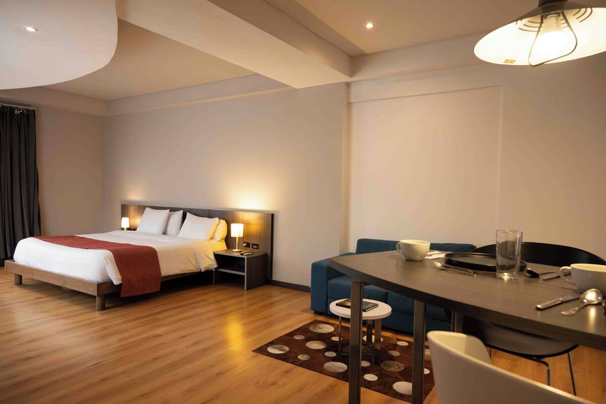 Ein Hotelzimmer mit Bett, Nachttisch, Lampe, Esstisch mit Stühlen und einem kleinen Hocker.