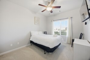 A bedroom with a bed, a ceiling fan, a window with curtains, and a desk.