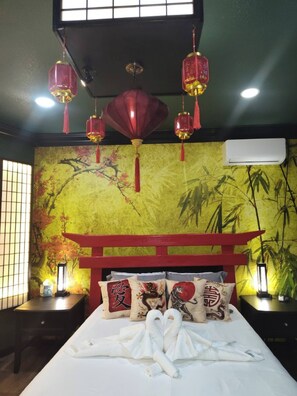 Une chambre avec un lit, une tête de lit rouge, des coussins décoratifs, des lanternes suspendues et un mur orné d’un tableau représentant des bambous.
