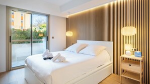 A modern hotel room with a large bed, a bedside table with a lamp, and a view of greenery through the window. - Terrasse vue Baie de Cannes - Welkeys (Le Cannet)
