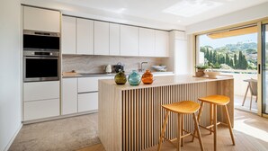 A modern kitchen with white cabinetry, a wooden island with stools, and a view of a hill through large windows. - Terrasse vue Baie de Cannes - Welkeys (Le Cannet)