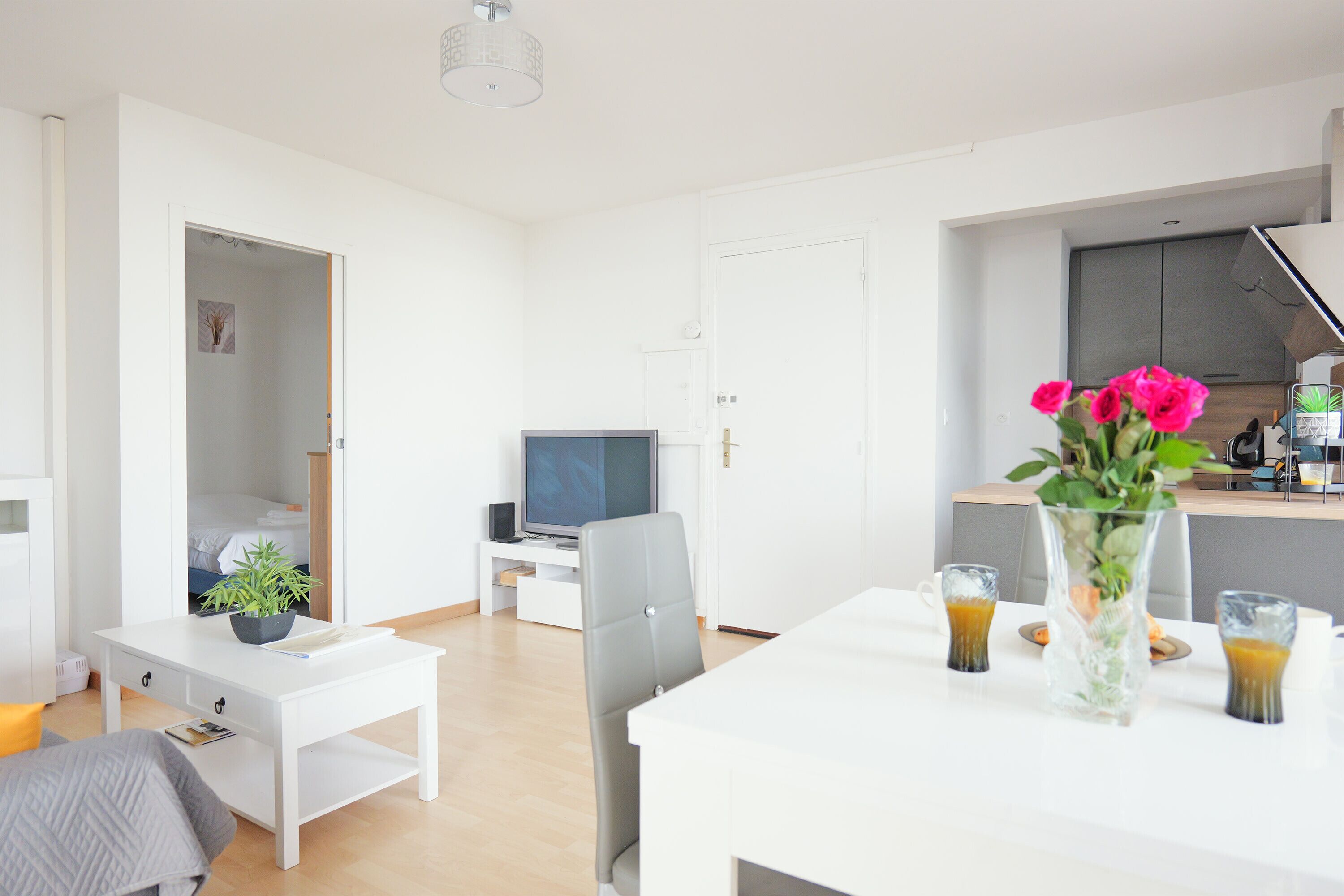 A modern living room with a white sofa, a small white table, a TV, and a kitchen area with a white countertop and a vase of flowers.