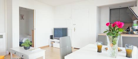 A modern living room with a white sofa, a small white table, a TV, and a kitchen area with a white countertop and a vase of flowers.