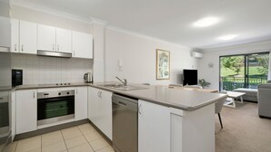 Una cocina moderna con armarios blancos, horno empotrado, lavavajillas y una isla grande con fregadero. Hay una zona de comedor con un sofá gris y un televisor.