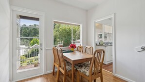 A dining area with a wooden table and chairs, a window with a view of greenery, and a kitchen visible through an open door. - Misty Pines in Leura (Leura)