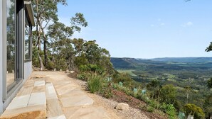 A stone pathway leading to a modern building with large windows, surrounded by lush greenery and a scenic view of rolling hills. - Sunset Retreat in Mount Victoria (Mount Victoria)
