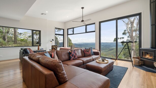 A modern living room with a leather sectional sofa, large windows offering a view of the outdoors, a ceiling fan, and a wood-burning stove. - Sunset Retreat in Mount Victoria (Mount Victoria)