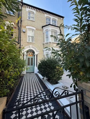A building with a checkered pathway leading to a green door, surrounded by greenery and a brick facade.