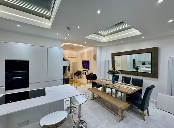 A modern kitchen with a dining area, featuring a built-in oven, a dining table with chairs, and a large mirror above the table.