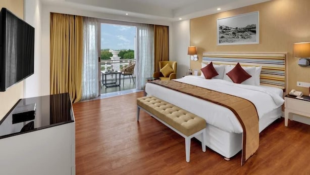 A hotel room with a large bed, a bench, a desk, a TV, and a balcony with a view. - Amanora the Fern Hotels And Club (Pune)