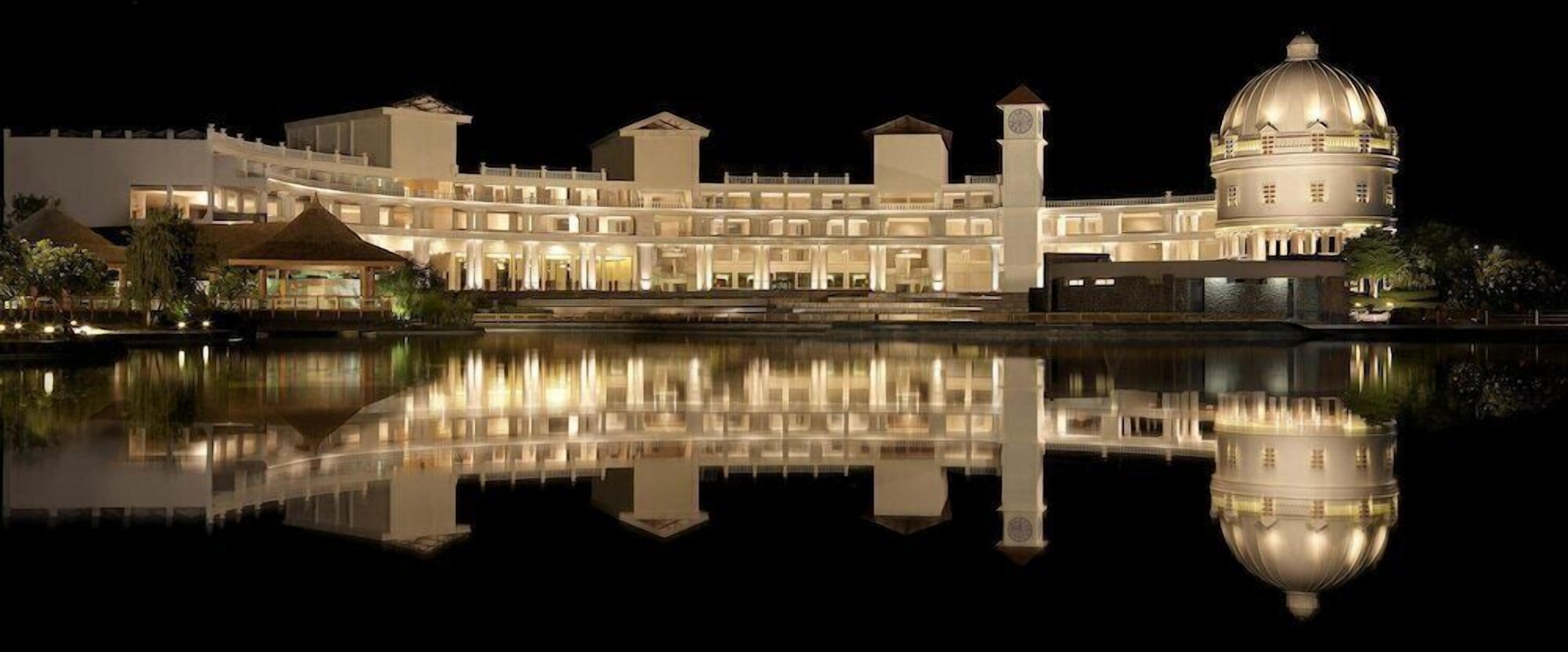 A large, multi-story hotel with a prominent dome and a clock tower, reflecting in a calm body of water.