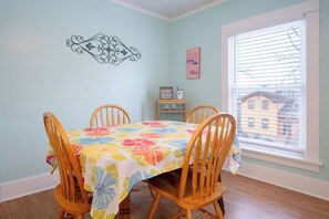 Un coin repas avec une table recouverte d’une nappe à motifs floraux, entourée de chaises en bois. Un élément décoratif orne le mur au-dessus de la table, et une fenêtre avec des stores se trouve à droite.