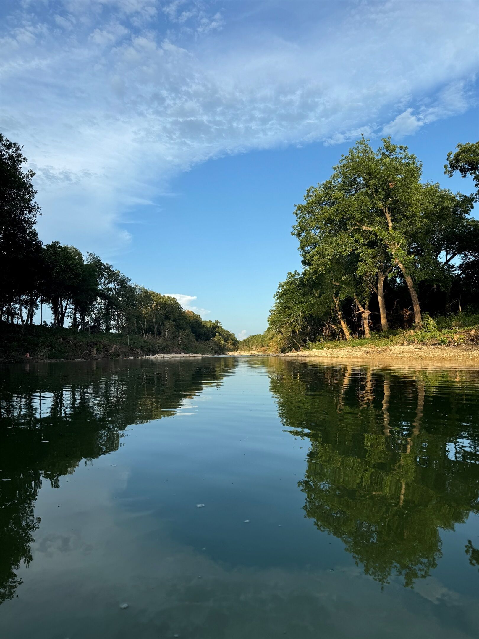 A calm river with trees on both sides, clear sky, and calm water reflecting the surroundings.