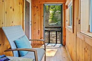 L’intérieur d’un chalet en bois, avec un fauteuil en osier, une fenêtre donnant sur une voiture et des arbres, et un balcon doté d’une balustrade.