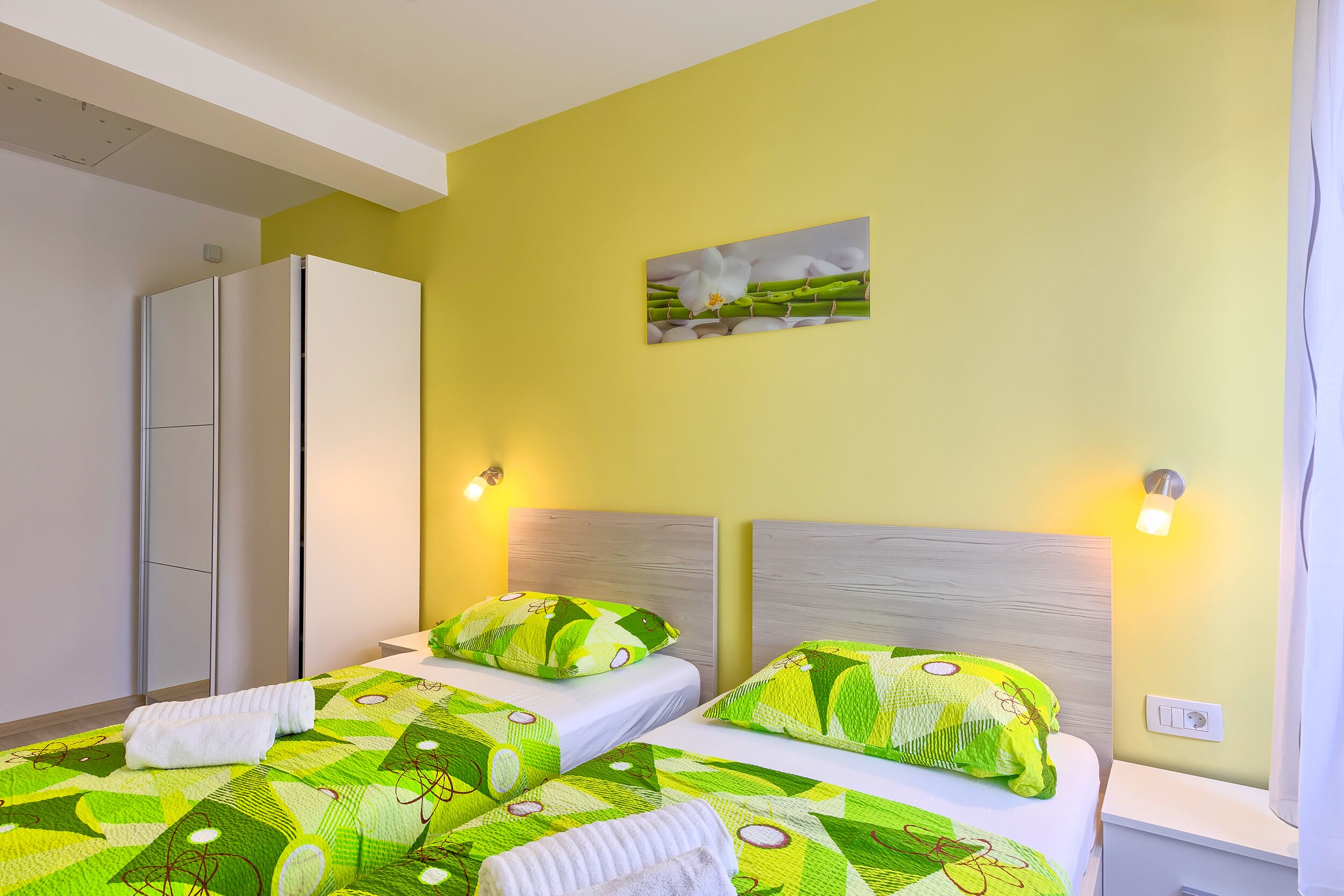A hotel room with two beds, each with green patterned bedding and white pillows, a painting on the wall, and a wardrobe.