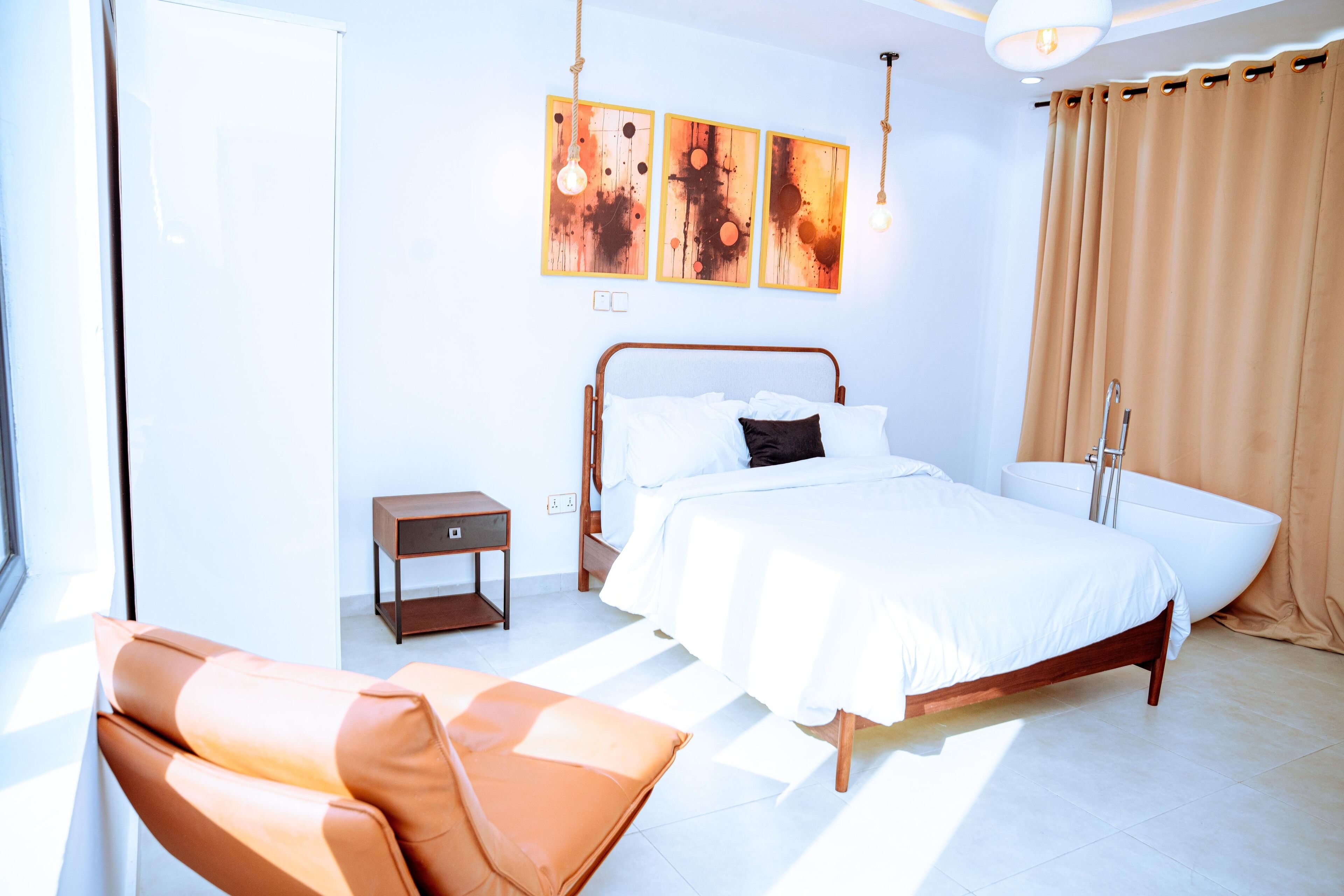A modern bedroom with a bed, a side table, a chair, a bathtub, and artwork on the wall.