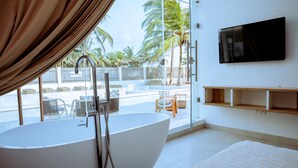 A modern bathroom with a large bathtub, a TV mounted on the wall, and a view of the pool area. - Atlantic Shores Resort (Lagos)